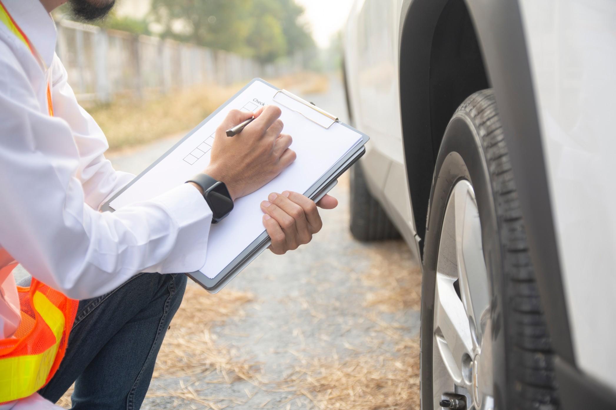 A person kneeling next to a car with a clipboard doing a safety inspection.