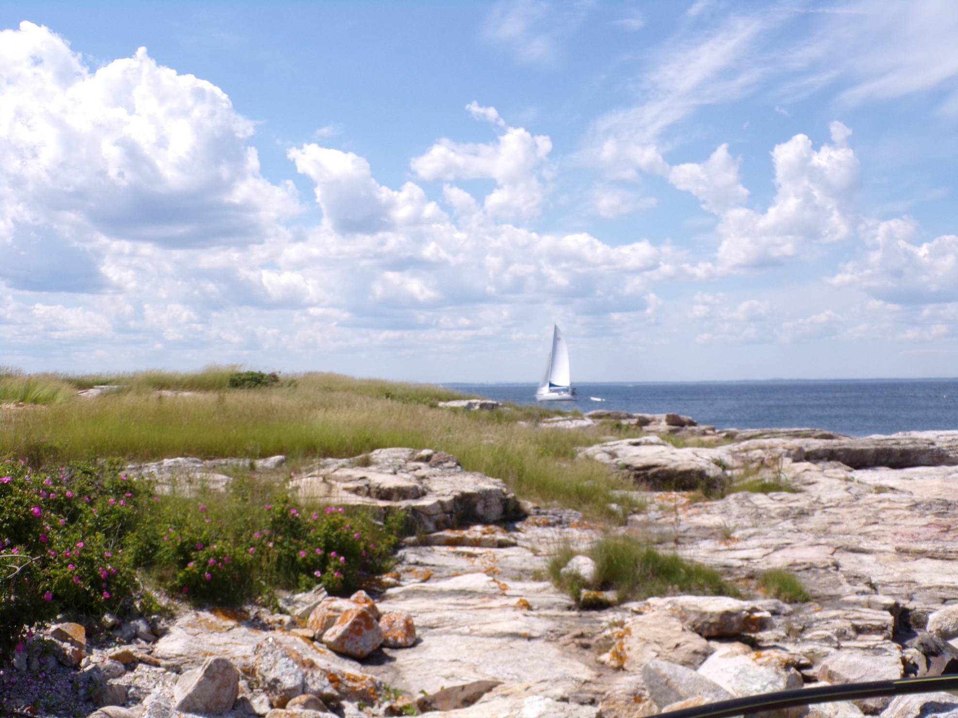 The View from Star Island, Isles of Shoals in New Hampshire. Blue cloudy sky, rocky coast, and a white sailboat on the sea. 