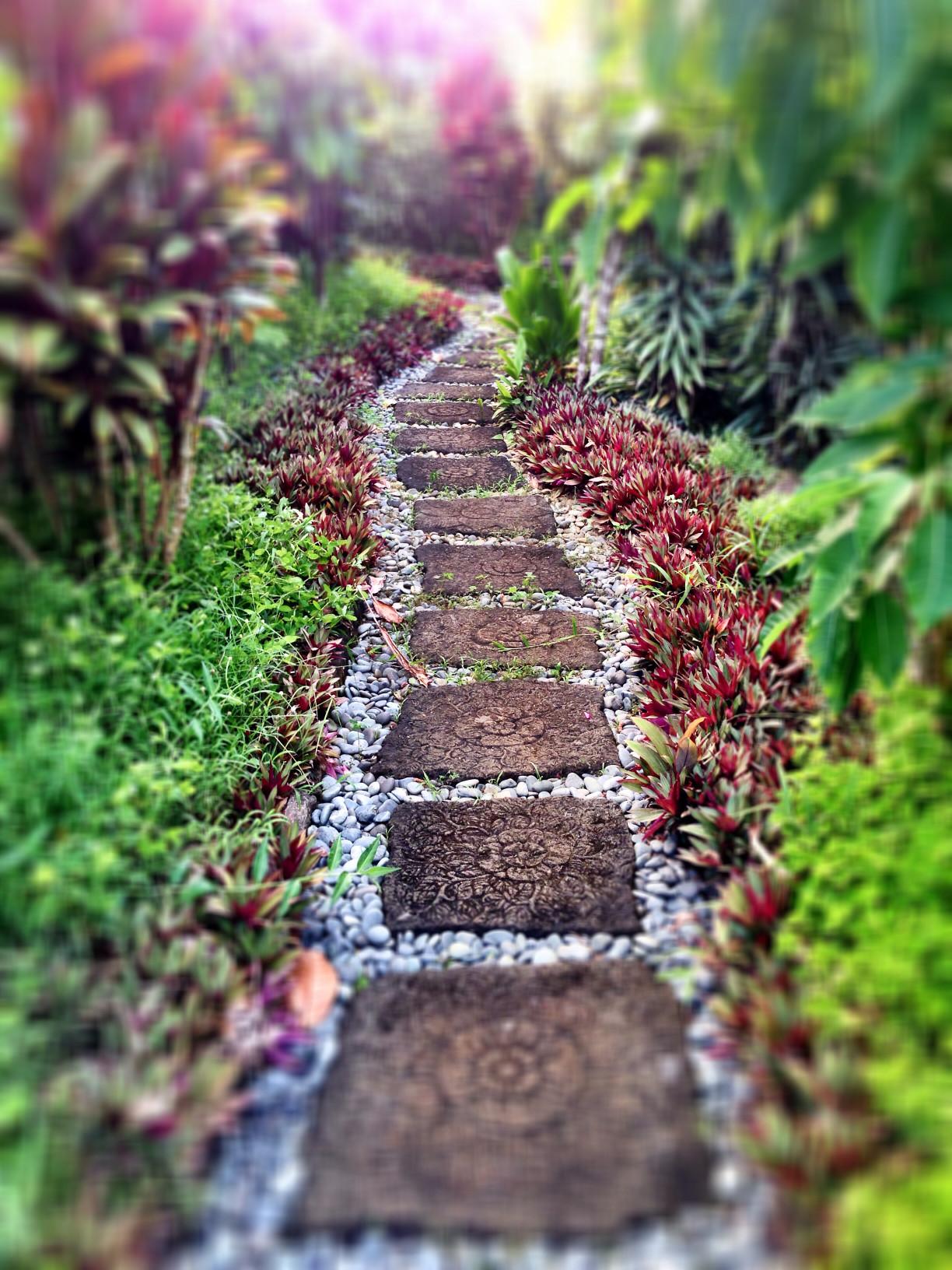 White and gray pebble path with brown flat stones set on top, lush garden surrounding.