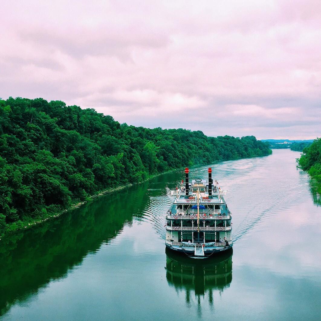 Ferry boat moving down the river towards the viewer under a cloudy sky with woods on either bank. 